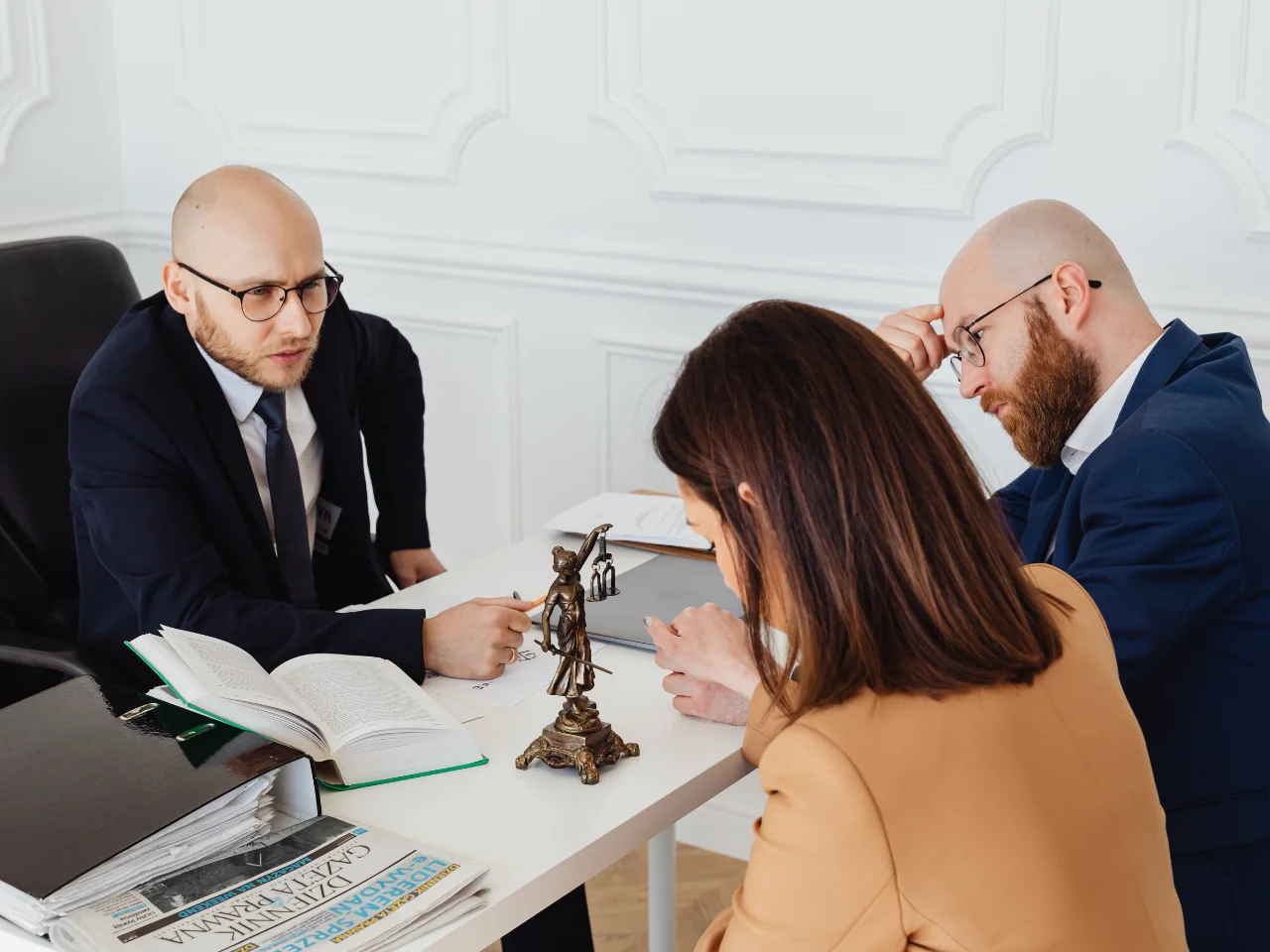 Three people in formal attire sit around a desk with legal books, papers, and a Lady Justice statue, appearing to discuss something seriously in an office setting.