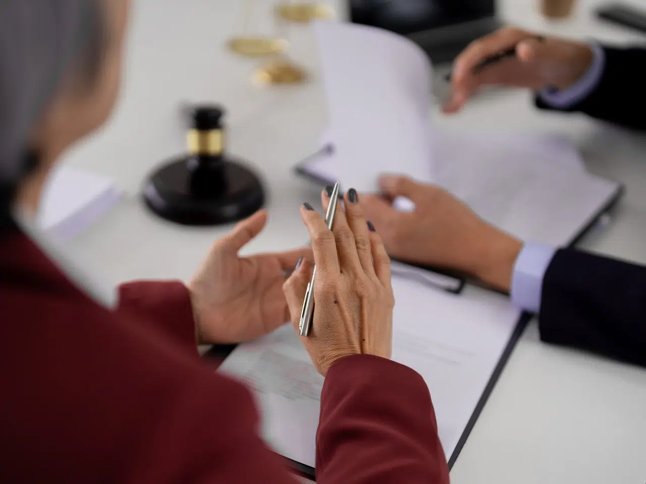 Two people in business attire sit at a desk with documents, one holding a pen. A gavel is visible on the table, suggesting a legal or judicial discussion or consultation.