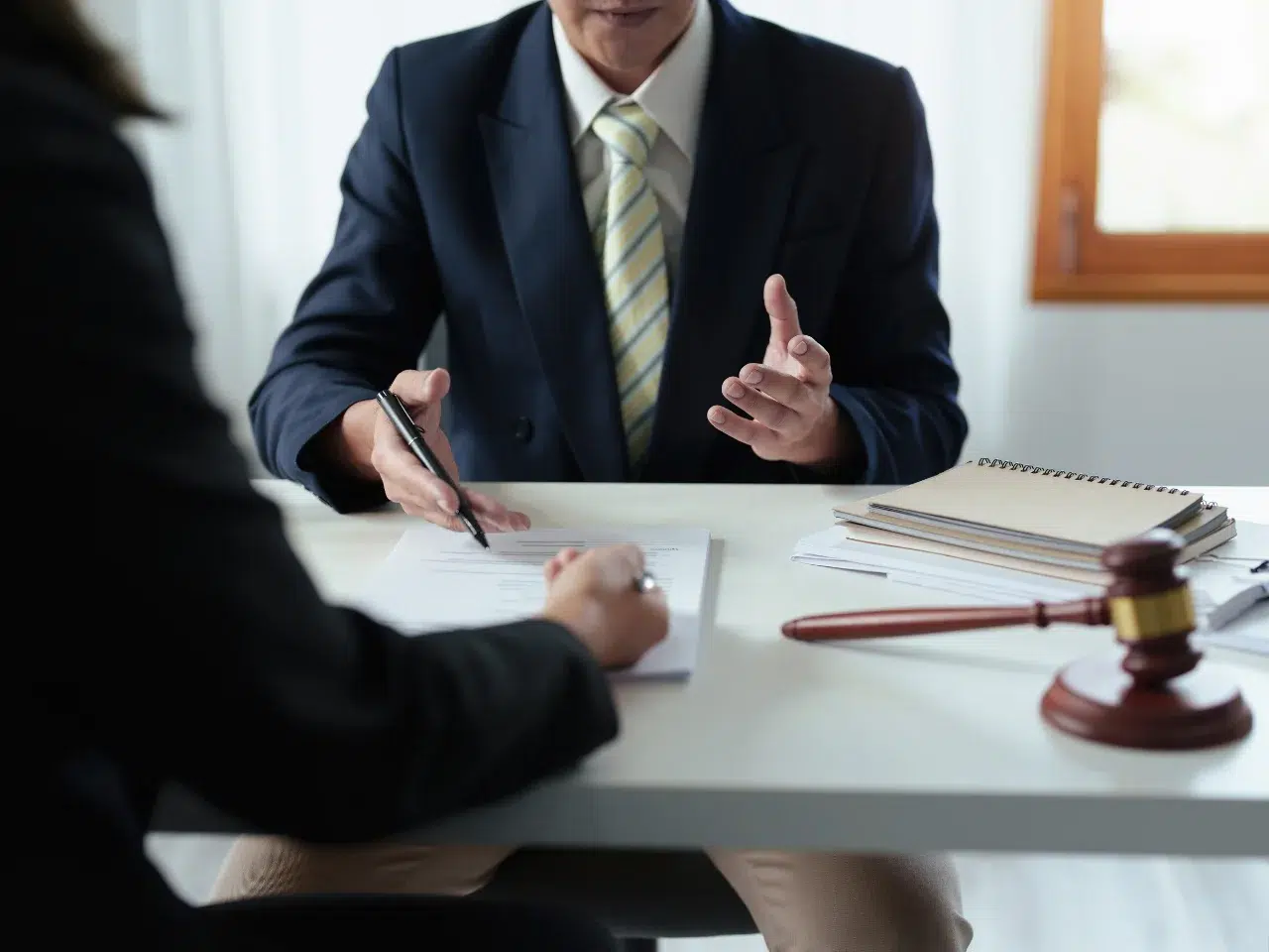 Two people sit at a desk discussing documents; one holds a pen, and the other gestures with hands. A gavel and notebooks are on the table, suggesting a legal or professional meeting.