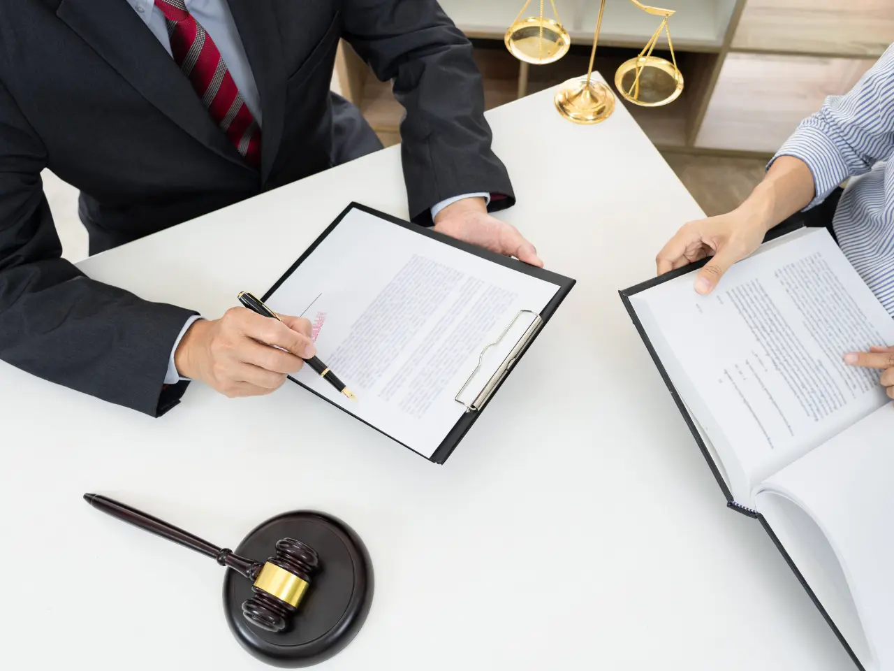 Two people sit at a desk reviewing documents on clipboards; one holds a pen pointing at the paper. A judges gavel and gold scales of justice are on the table, suggesting a legal or business meeting.