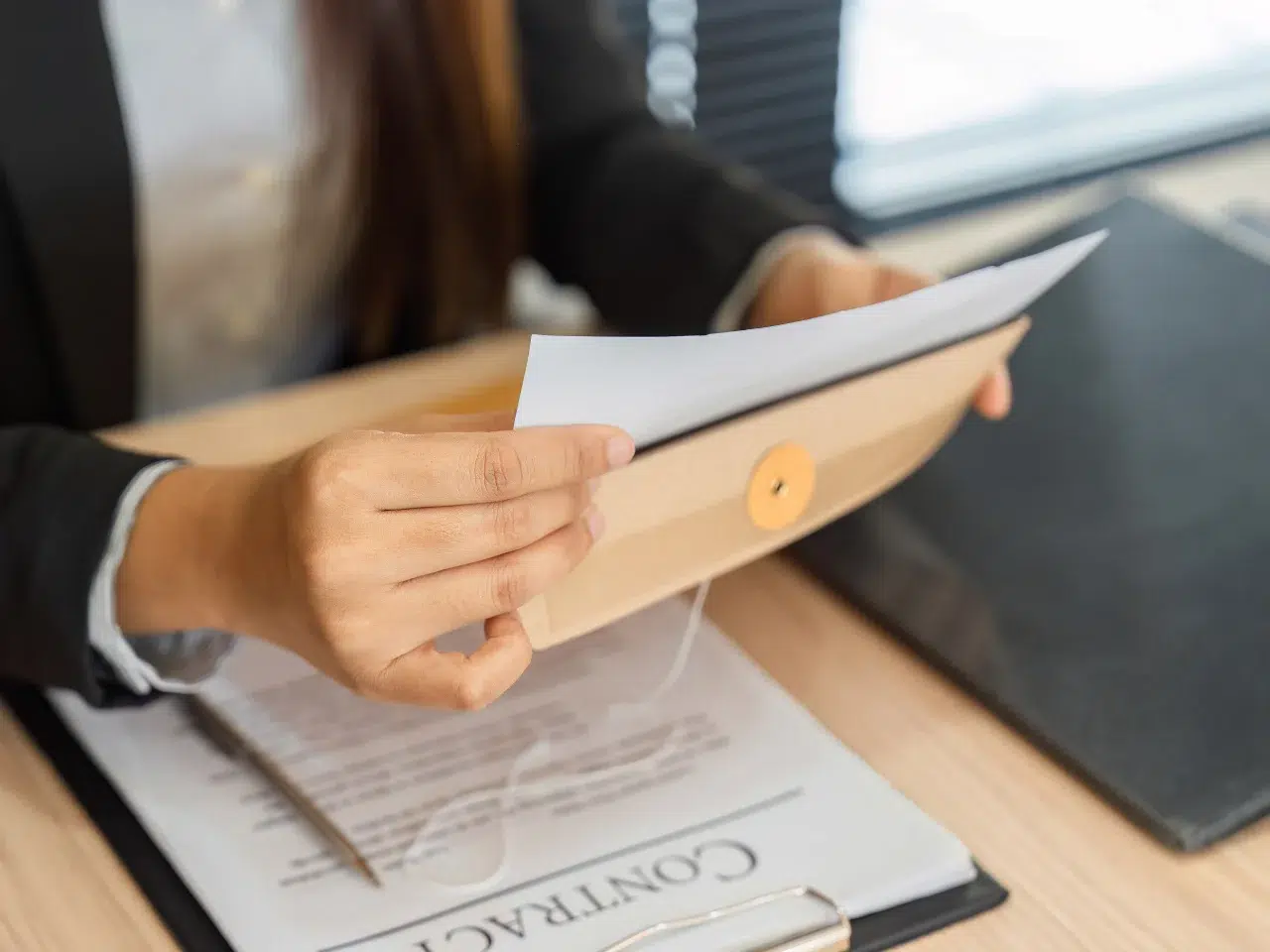 A person in business attire holding paperwork and a brown envelope, with a contract document visible on the desk underneath, suggesting a professional or legal setting.