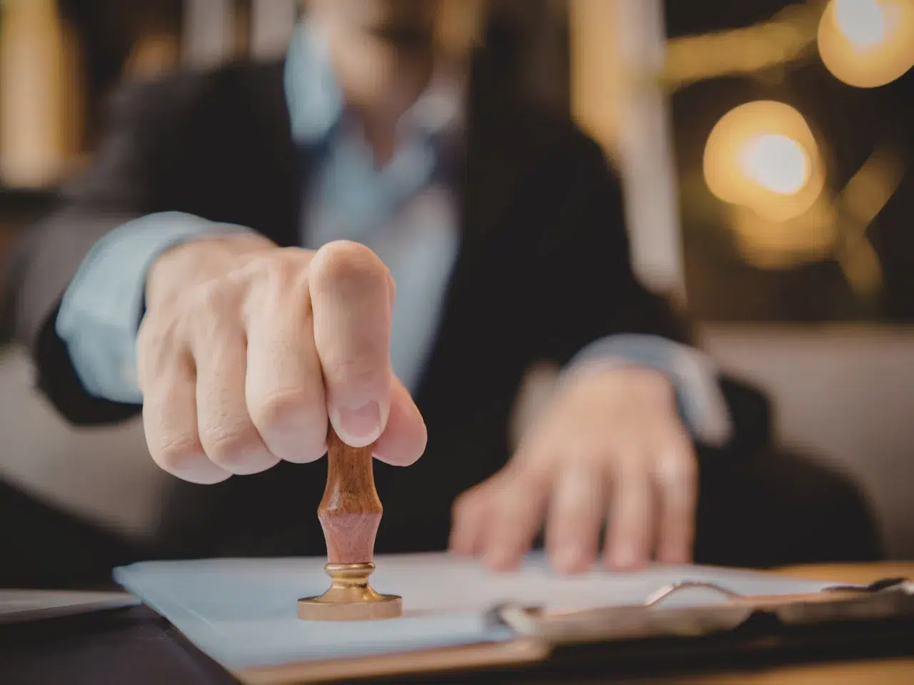 A person in a suit presses a wooden stamp onto a document on a desk, indicating approval or authentication. The focus is on the hand and stamp, with the face blurred in the background.