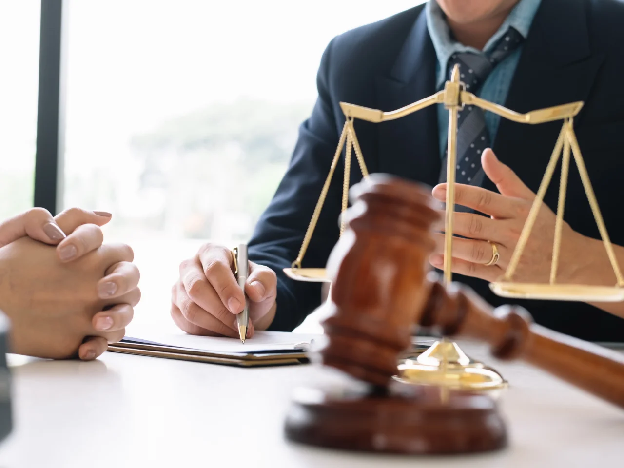 Two people sitting at a desk with a gavel, legal documents, and scales of justice; one person is taking notes while the other listens, suggesting a legal or consultation setting.