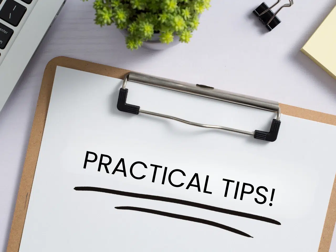 A clipboard with a paper that reads PRACTICAL TIPS! in bold letters, placed on a desk near a small green plant, a laptop corner, paper clips, and sticky notes.