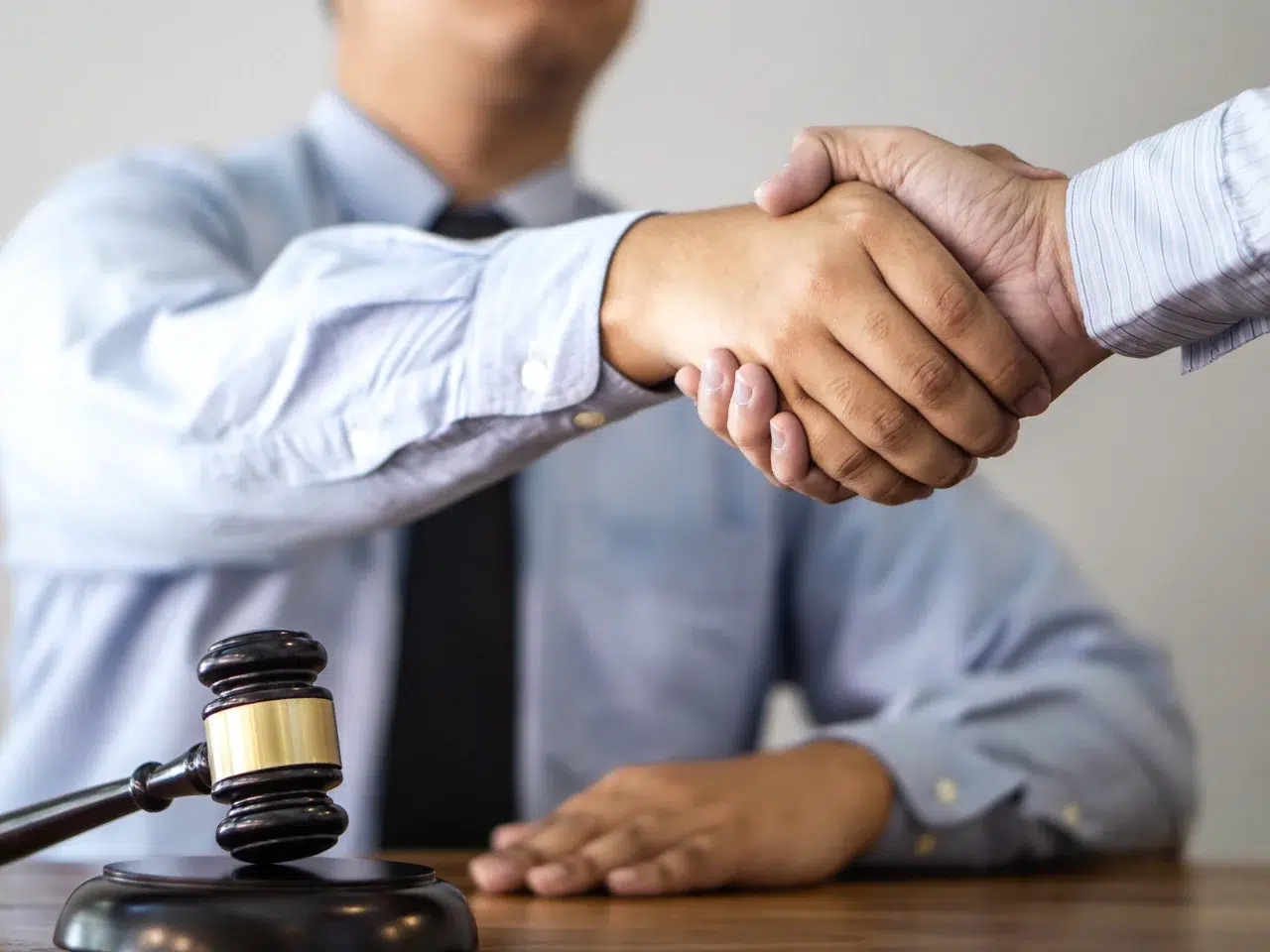 Two people in business attire shake hands across a desk with a gavel in the foreground, suggesting a legal agreement or settlement.