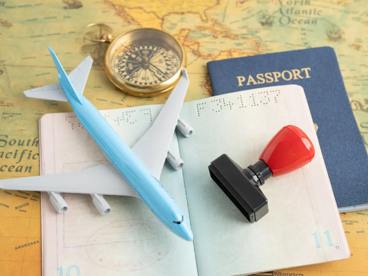 A toy airplane, a red stamp, and a gold compass rest on an open passport, with a blue passport booklet on an old world map, symbolizing travel and adventure.