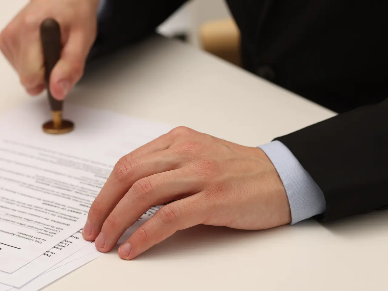 A person in a suit presses a stamp onto a document on a white desk, suggesting the approval or authorization of official paperwork.