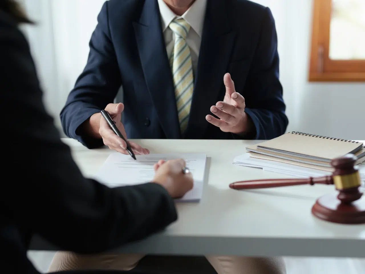Two people sit at a desk discussing documents; one is holding a pen, while the other gestures with their hands. A stack of notebooks and a judge’s gavel are visible on the desk, suggesting a legal or professional setting.