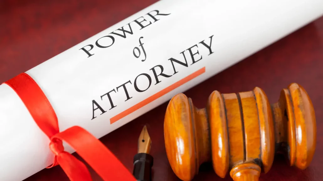 A scroll labeled Power of Attorney tied with a red ribbon lies next to a wooden judges gavel and a fountain pen on a reddish surface.