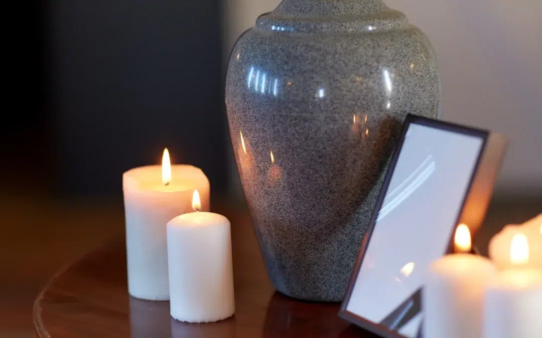 A gray urn sits on a wooden table next to three lit white candles and a picture frame with a blank or reflective surface.