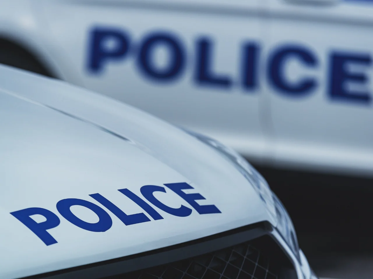 Close-up of a white police car hood with the word POLICE in bold blue letters, and another police vehicle with the same lettering blurred in the background.