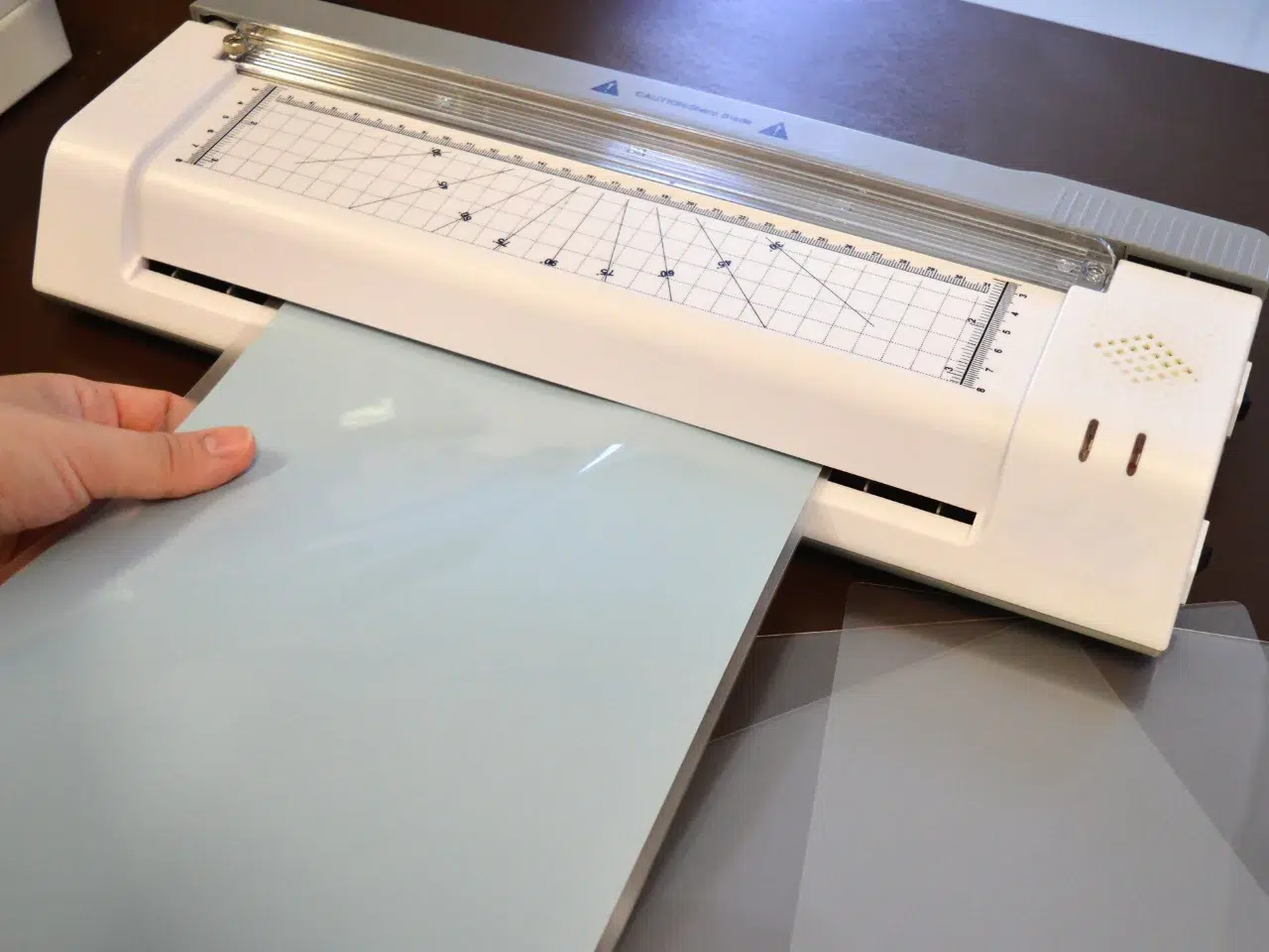 A hand is feeding a sheet of paper into a paper cutter with a grid and measurement markings, ready to trim the edge of the paper.