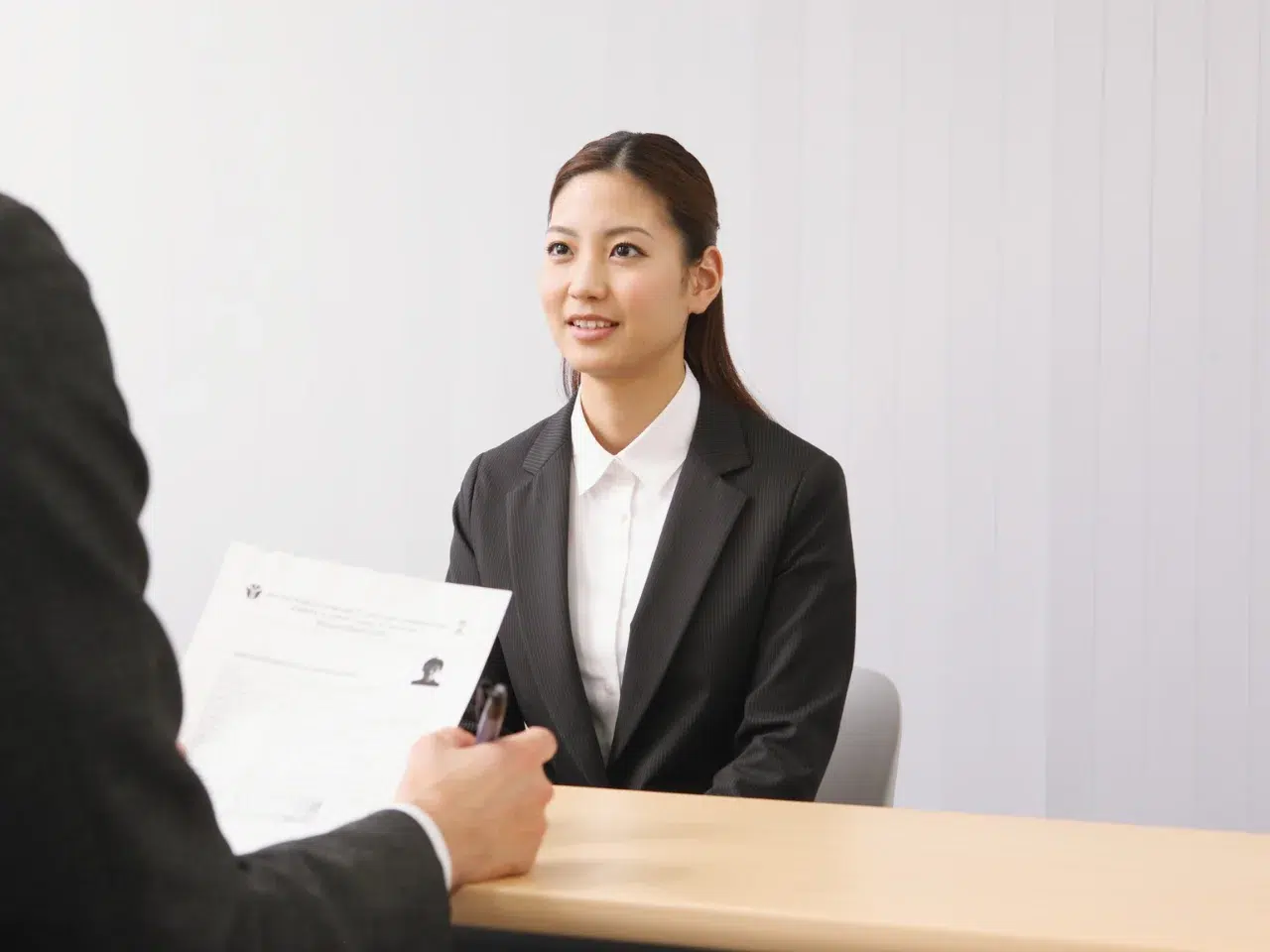 A woman in a black suit and white shirt sits at a desk, smiling during an interview. Across from her, a person holds a resume and a pen. The background is plain and light-colored.