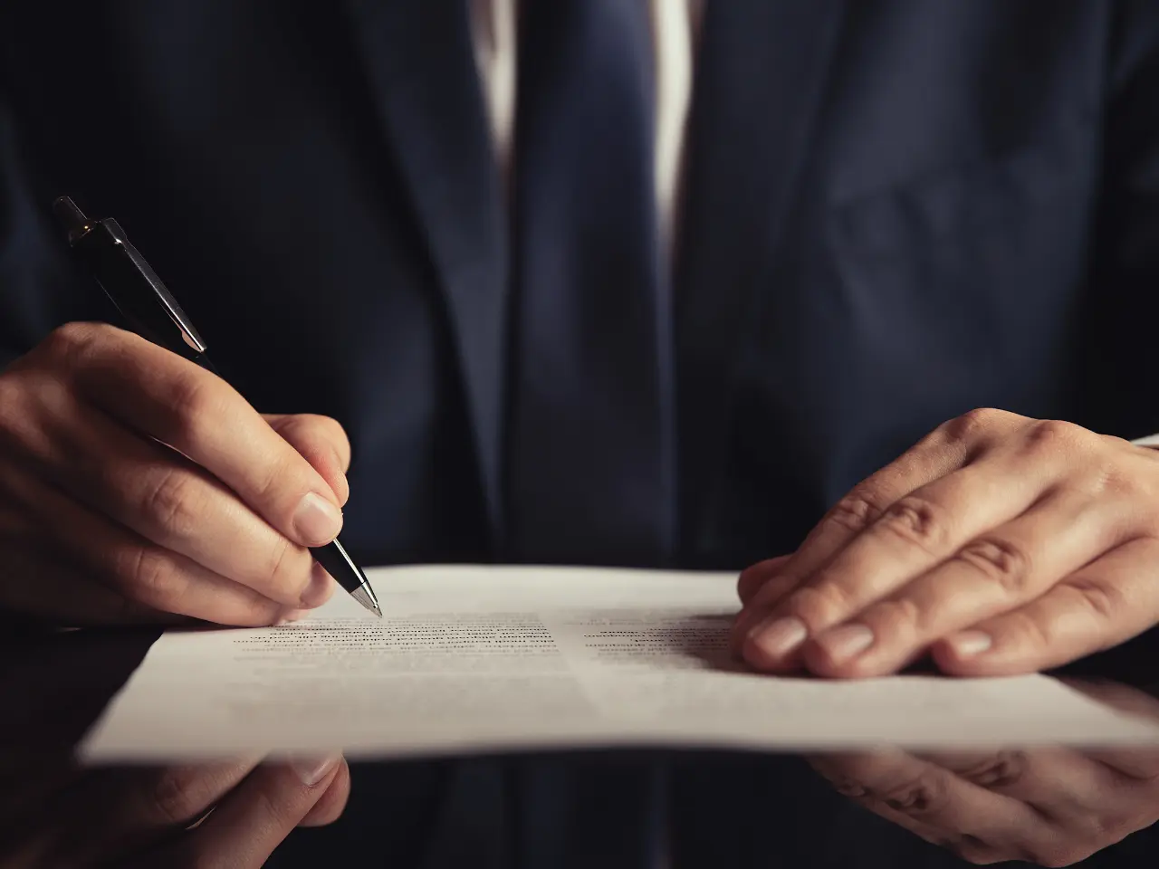 A person in a dark suit and tie is signing a document with a pen. Only their hands, pen, paper, and part of their suit are visible on a dark reflective surface.