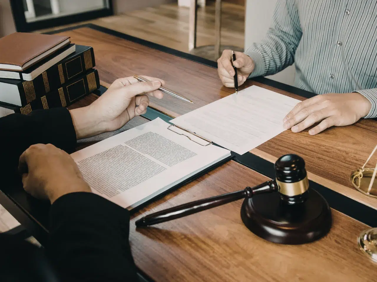 Two people sit at a wooden desk reviewing and signing legal documents. A gavel, legal books, and a clipboard with paperwork are on the desk, suggesting a legal or law office setting.