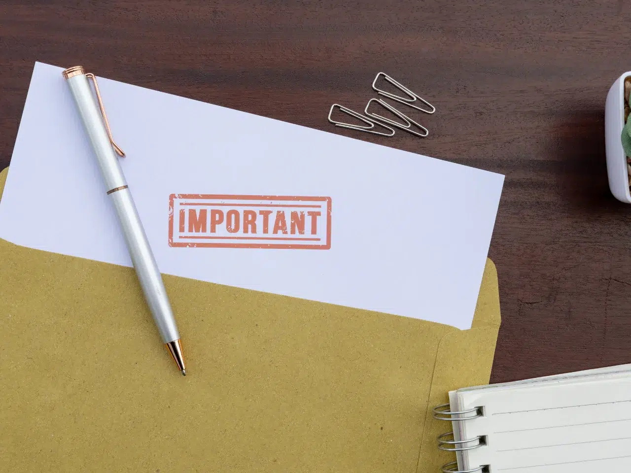 A white envelope stamped with IMPORTANT in red ink lies on a brown folder, next to a silver pen, three paperclips, and a lined notebook on a wooden desk.