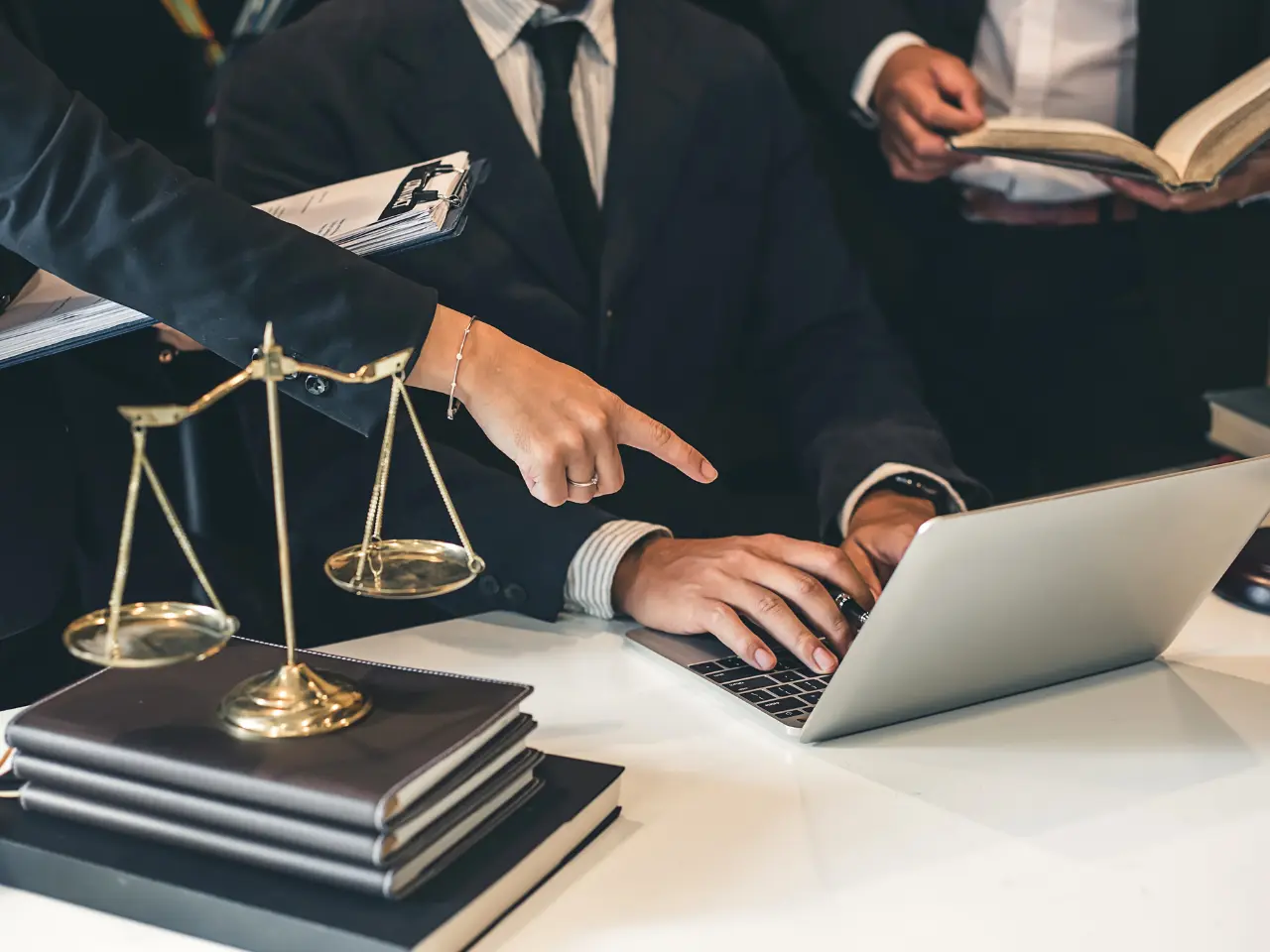 A person in a suit types on a laptop while another person points at the screen; legal scales, stacked books, and documents are on the desk, suggesting a professional or legal setting.