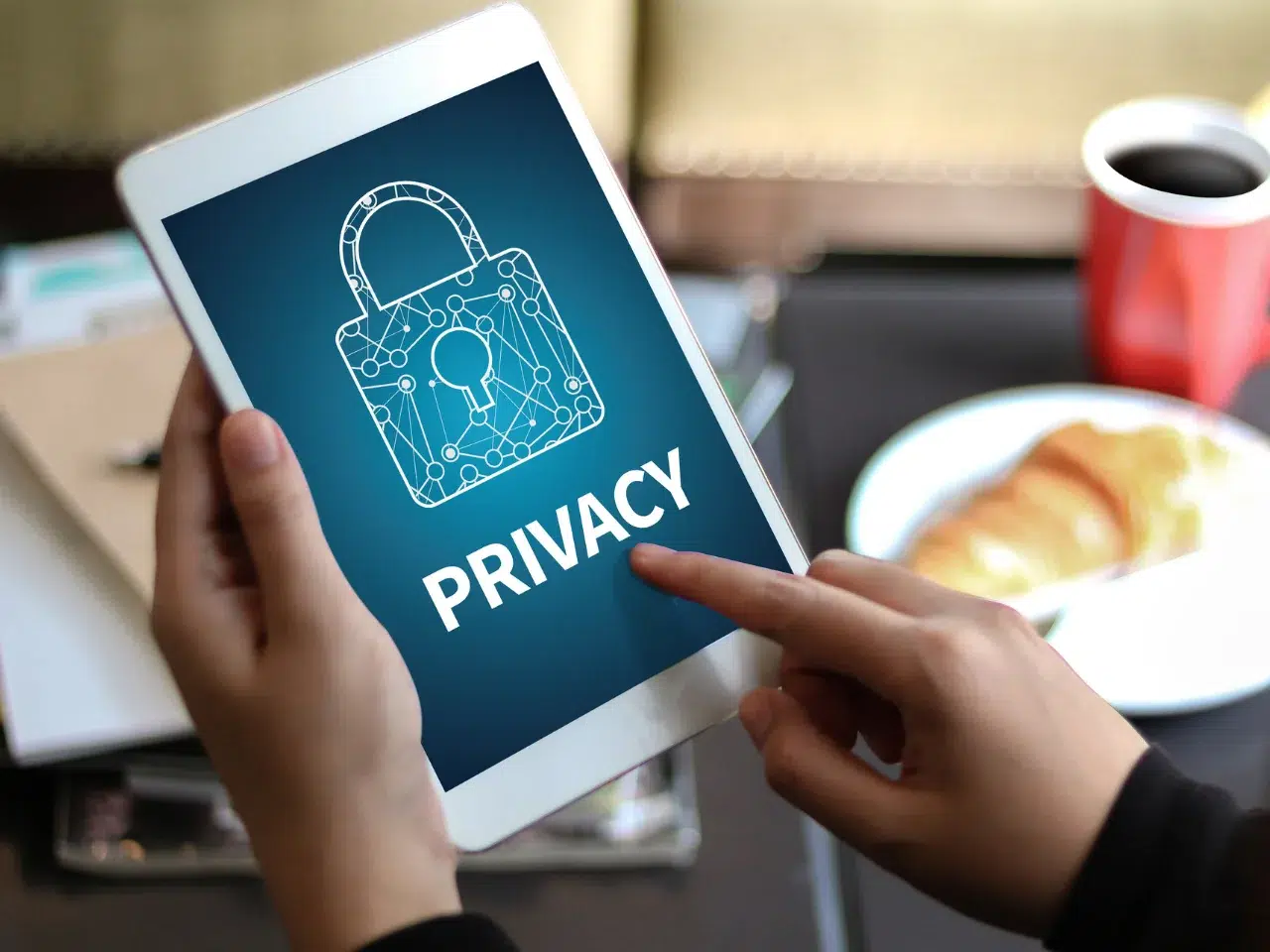 A person holds a tablet displaying an image of a digital padlock and the word “PRIVACY” on the screen. In the background, there is a plate of food and a cup of coffee on a table.