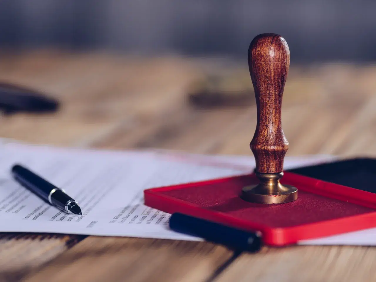 A wooden rubber stamp sits on a red ink pad next to a black pen and official-looking documents on a wooden table.
