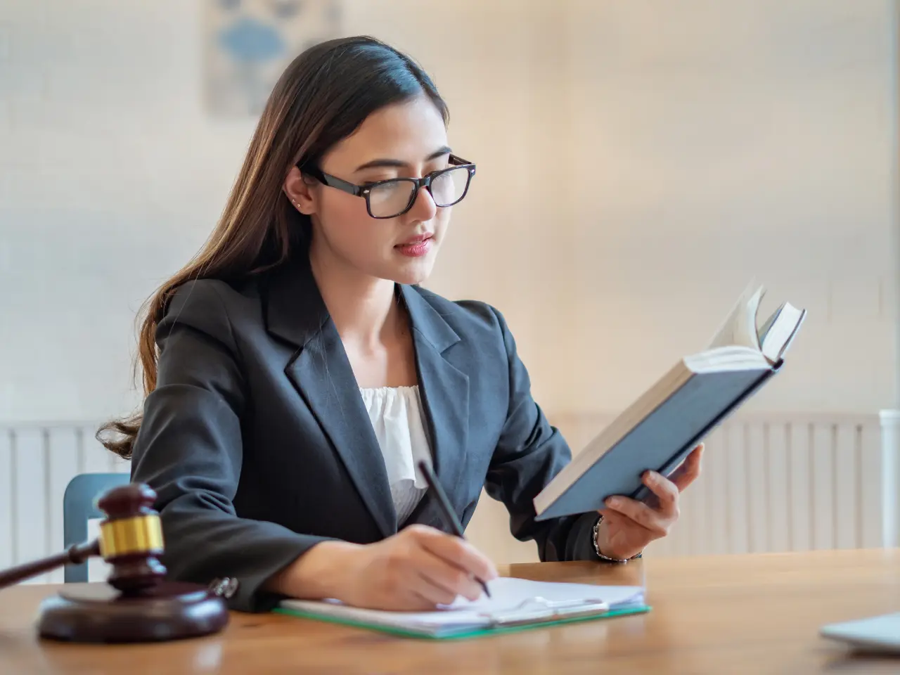 A woman in a blazer and glasses sits at a desk reading a book and taking notes. A gavel is visible on the desk, suggesting a legal or law-related setting.