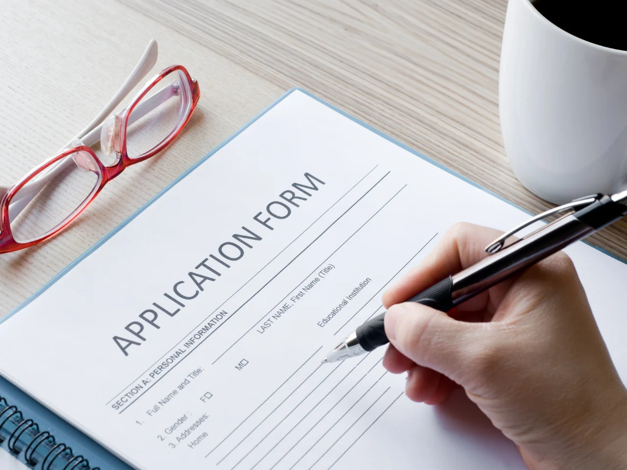 A person fills out an application form on a desk with a pen. Red eyeglasses, a white coffee cup, and part of a blue folder are also visible on the light wooden surface.