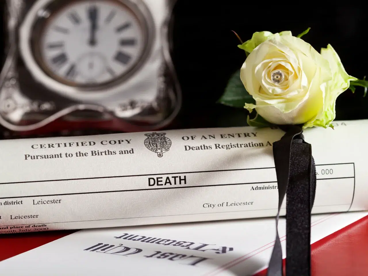 A death certificate rolled up with a white rose and a black ribbon on top, next to a document labeled Last Will & Testament and a blurred clock in the background.
