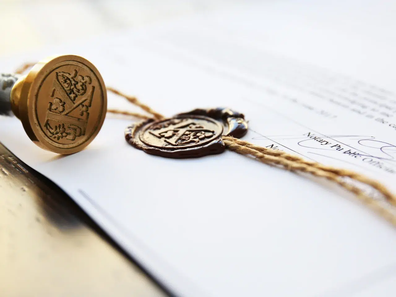A close-up of a document sealed with brown wax and twine, featuring an embossed metal stamp with ornate designs next to the seal. The document includes text and a partial handwritten signature.