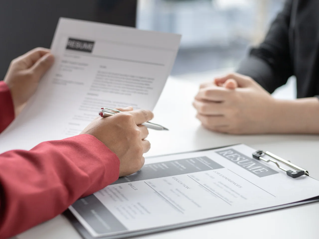 Two people sit across a table during a job interview; one holds a résumé and a pen, while a résumé on a clipboard lies on the table in front of the other person.