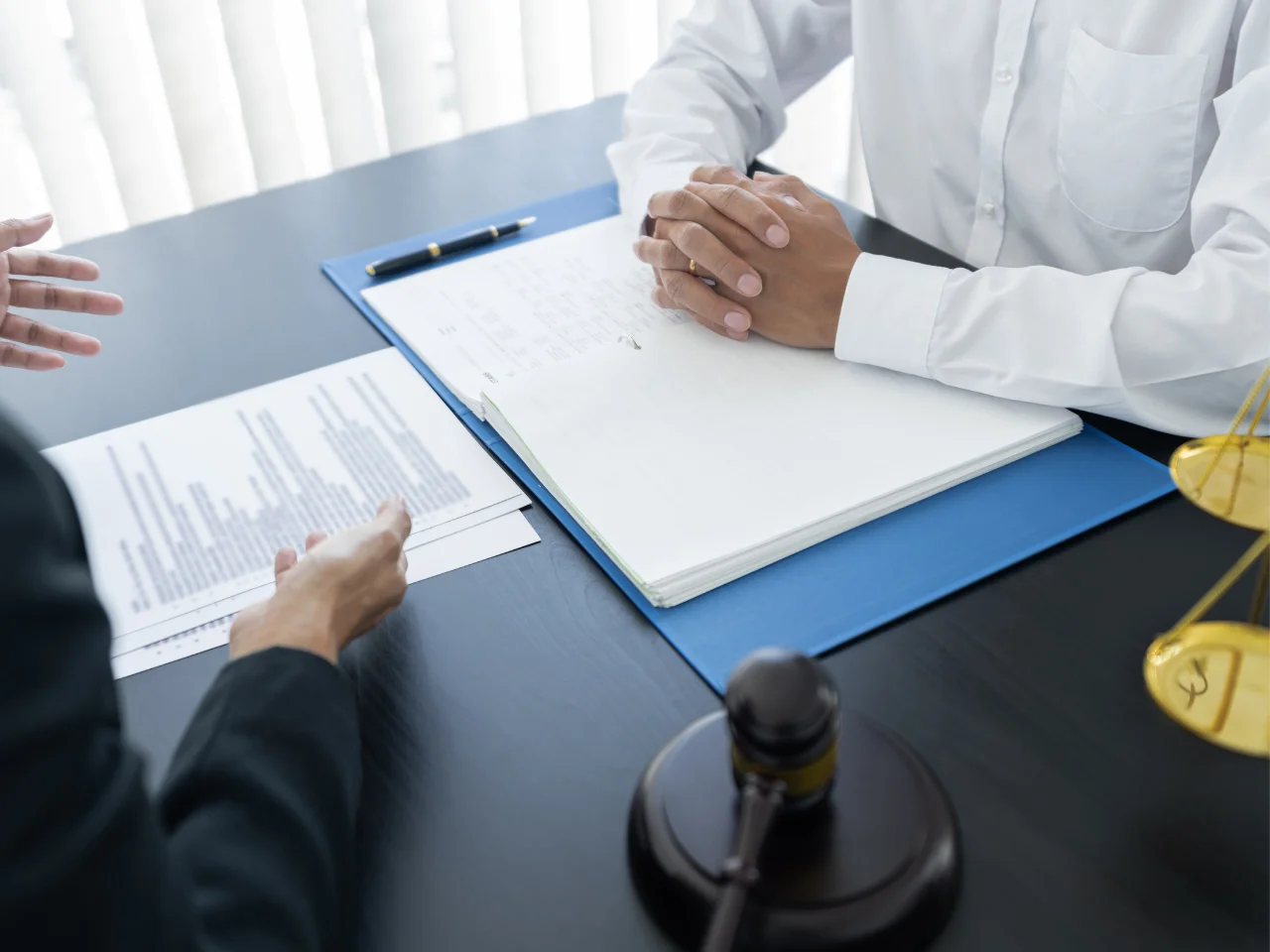 Two people sit at a desk with legal documents, a gavel, and scales of justice; one person gestures while the other listens with hands clasped, suggesting a legal consultation or discussion.