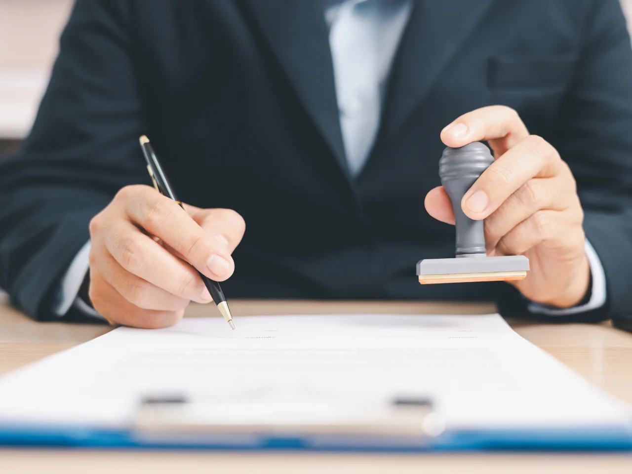 A person in a suit is holding a pen in one hand and a rubber stamp in the other, preparing to sign and stamp a document on a desk. The focus is on the hands and the paperwork.
