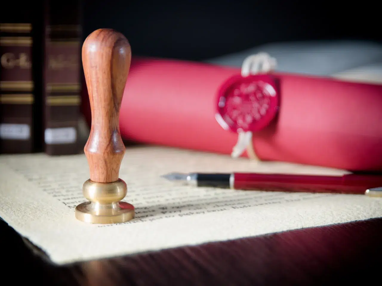 A wooden stamp, a fountain pen, and a rolled red document sealed with wax rest on a legal or official-looking paper, suggesting a scene of authentication or certification.