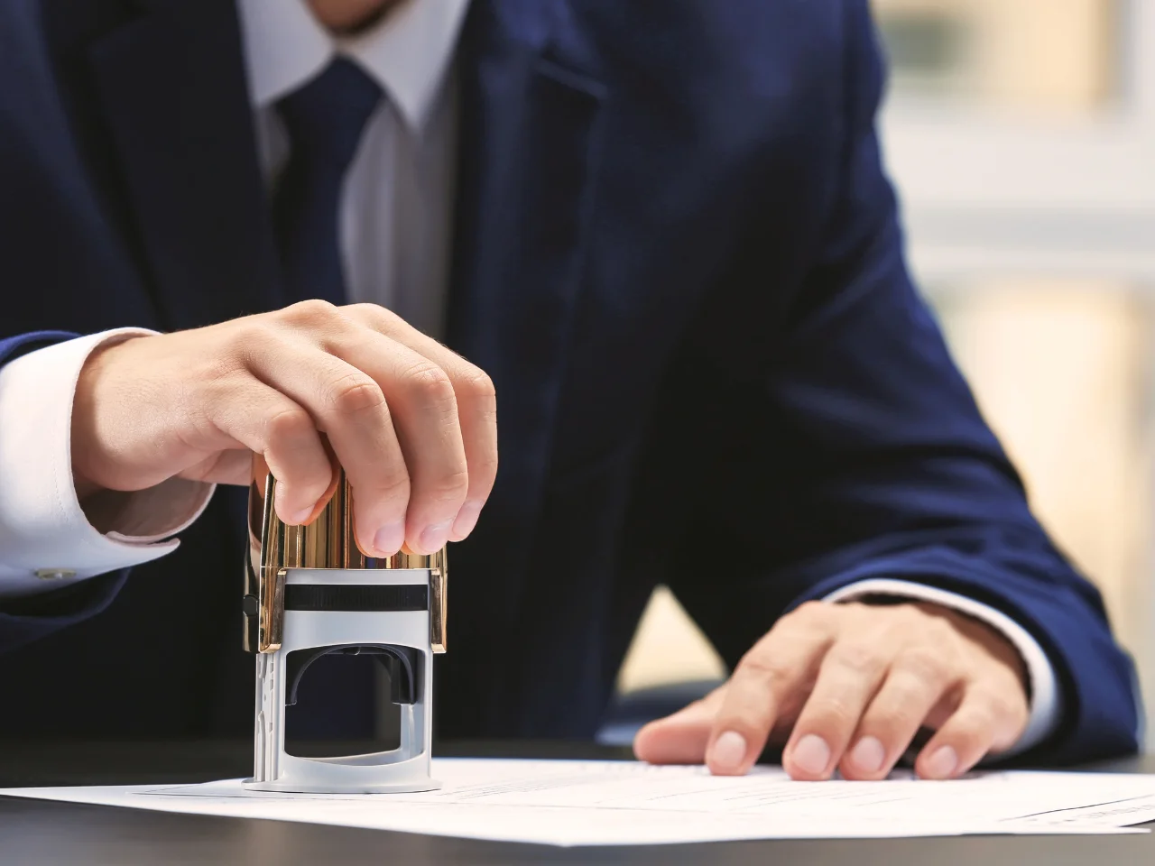 A person in a dark suit stamps an official document on a desk, using a metal and plastic stamp, with only their hands and torso visible.