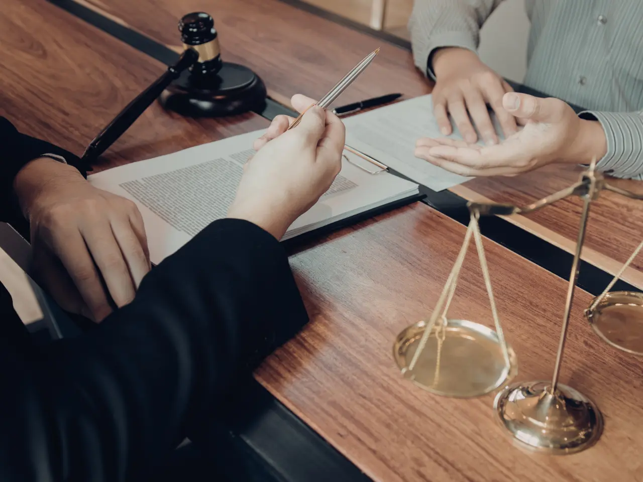 Two people sit at a desk with legal documents; one holds a pen while the other gestures with open hands. A gavel and a set of scales are visible, suggesting a legal or courtroom setting.
