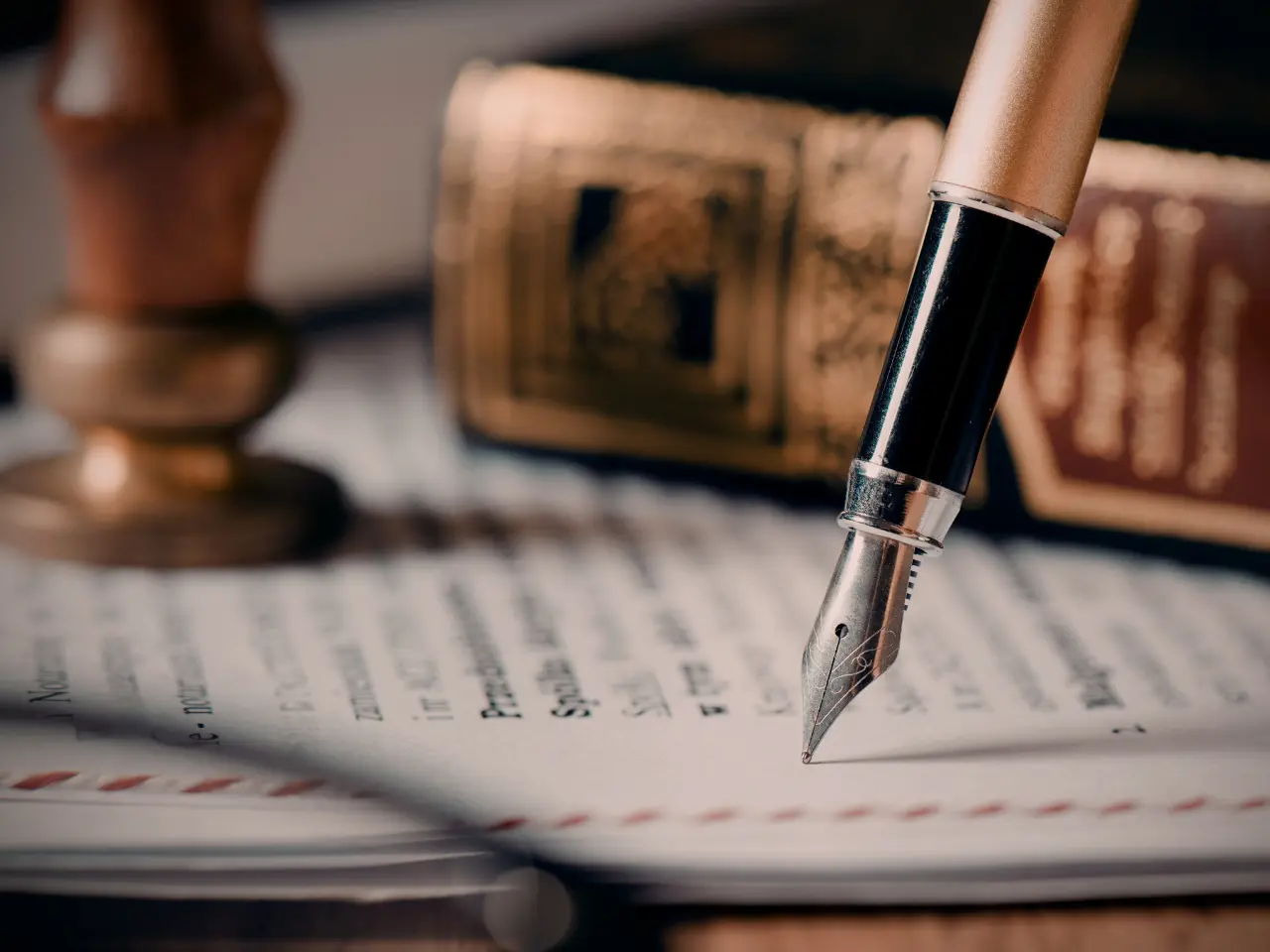 A close-up of a fountain pen resting on a document, with a blurred book and a wax seal stamp in the background, suggesting a classic writing or legal theme.