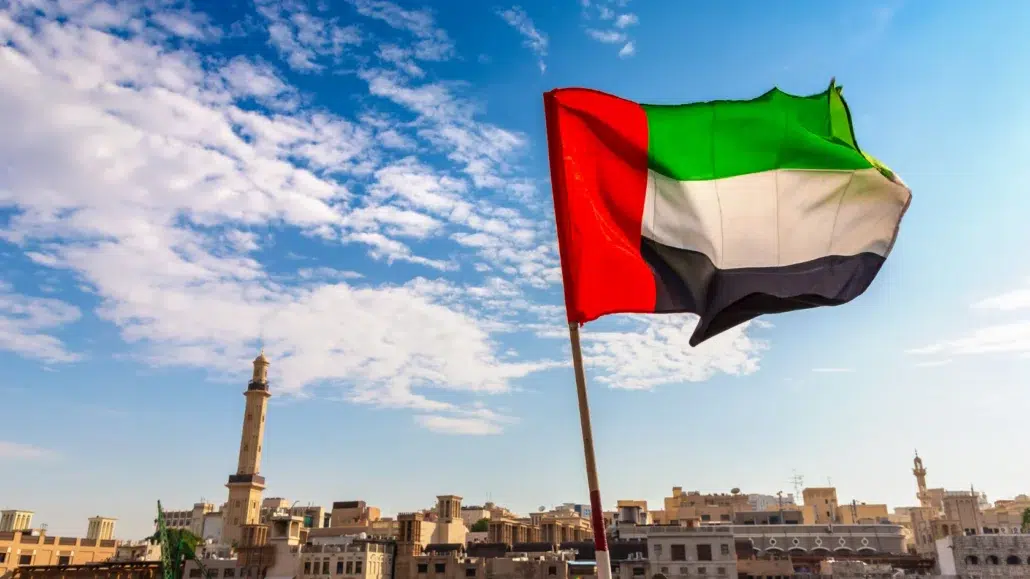 The flag of the United Arab Emirates waves against a blue sky with clouds, overlooking a cityscape with traditional buildings and a tall minaret in the background.
