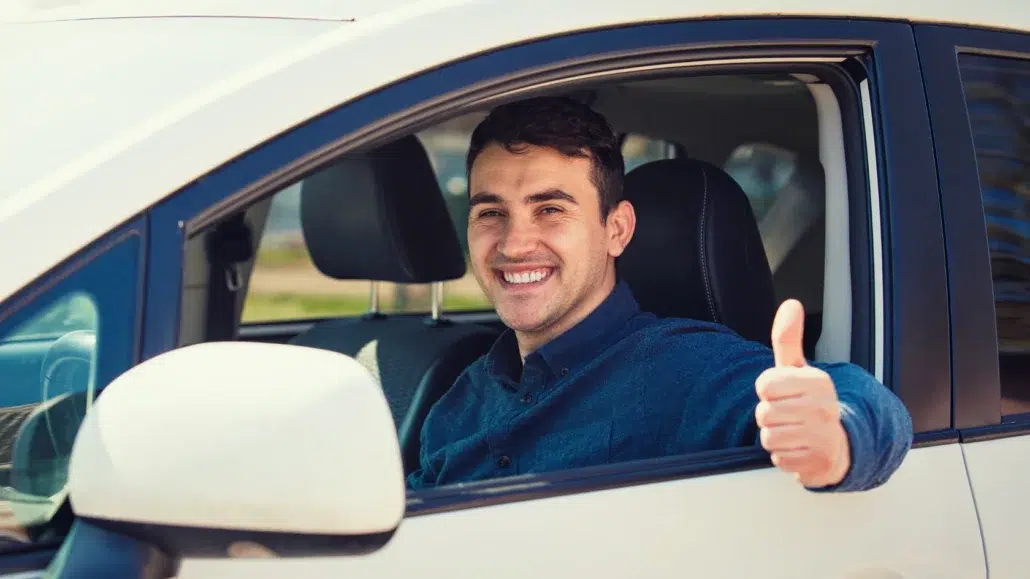 A smiling man sits in the drivers seat of a white car with the window down, giving a thumbs-up gesture to the camera.