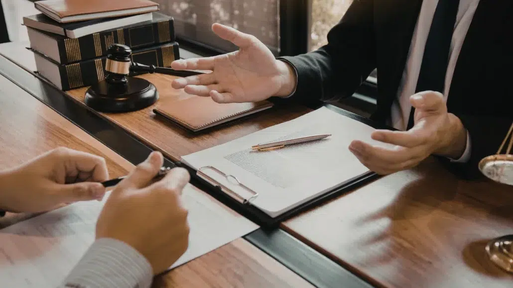 Two people sit at a desk discussing legal documents. One person gestures with their hands while the other holds a pen. A gavel, books, and paperwork are on the desk, suggesting a legal or consultation setting.