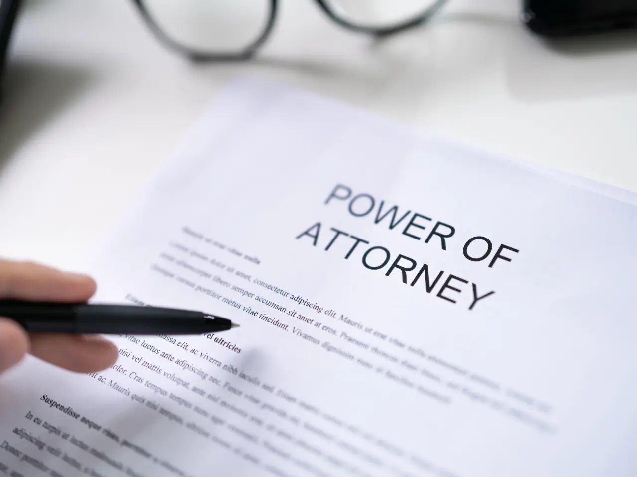 A close-up of a Power of Attorney document on a table, with a hand holding a black pen pointing at the paper. A pair of eyeglasses is also visible in the background.