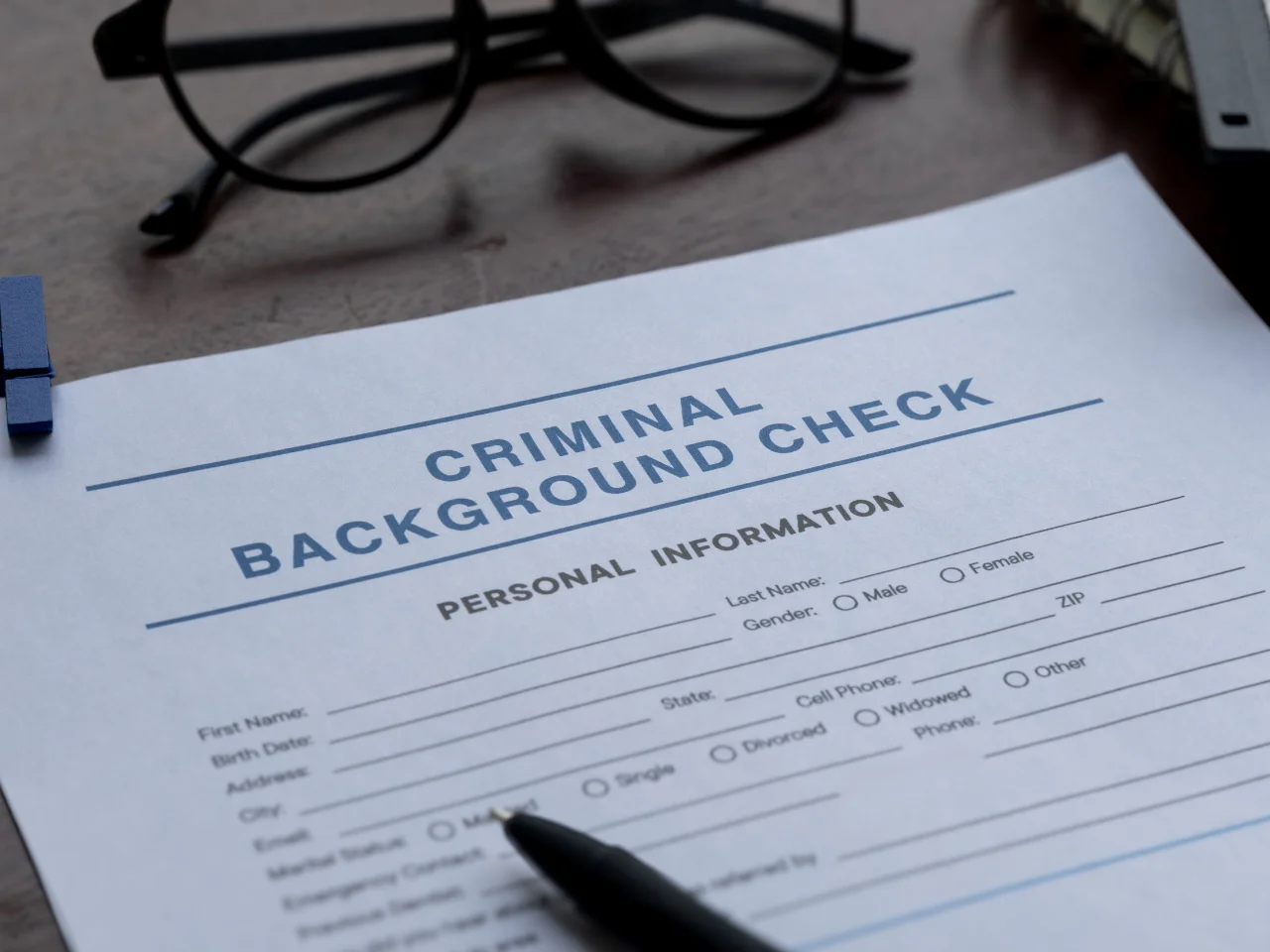 A close-up of a criminal background check form with sections for personal information. A black pen rests on the form, and a pair of eyeglasses is visible in the background.