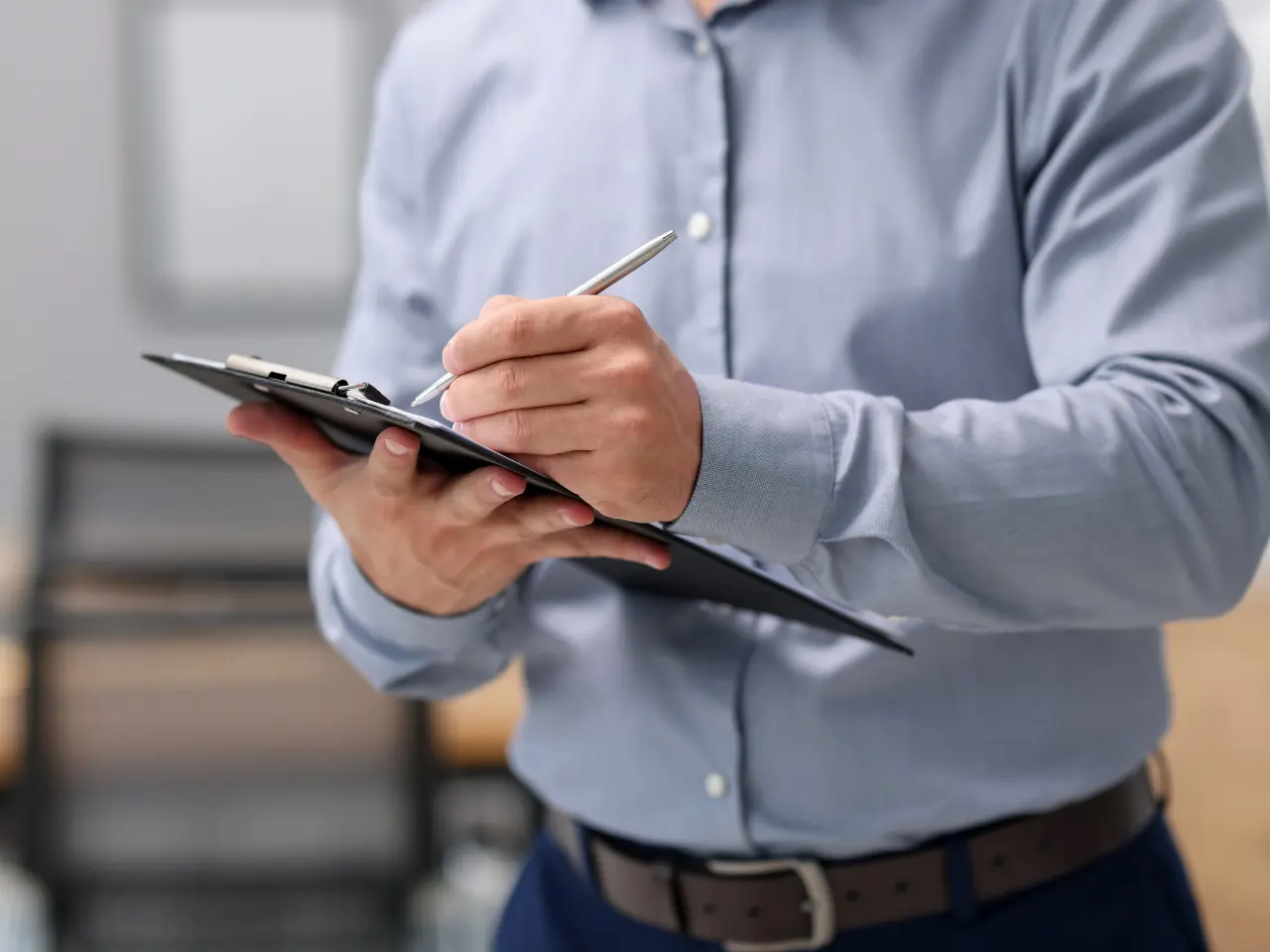 A person wearing a light blue dress shirt and brown belt is holding a clipboard and writing with a pen. The background is blurred, focusing on the person’s torso and hands.