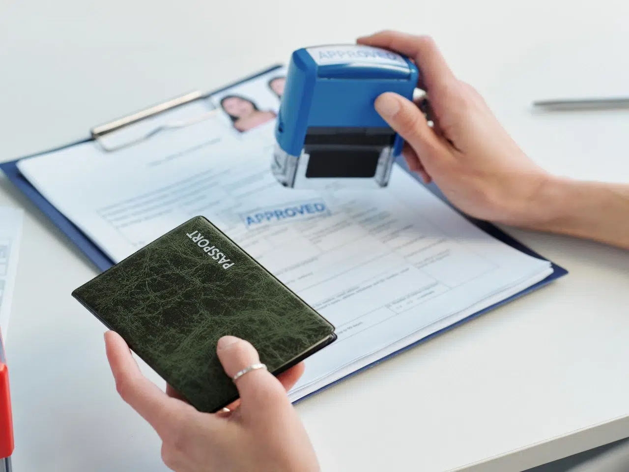 A person holds a green passport and uses a blue APPROVED stamp on a document with a photo, sitting on a clipboard, indicating official approval or processing.