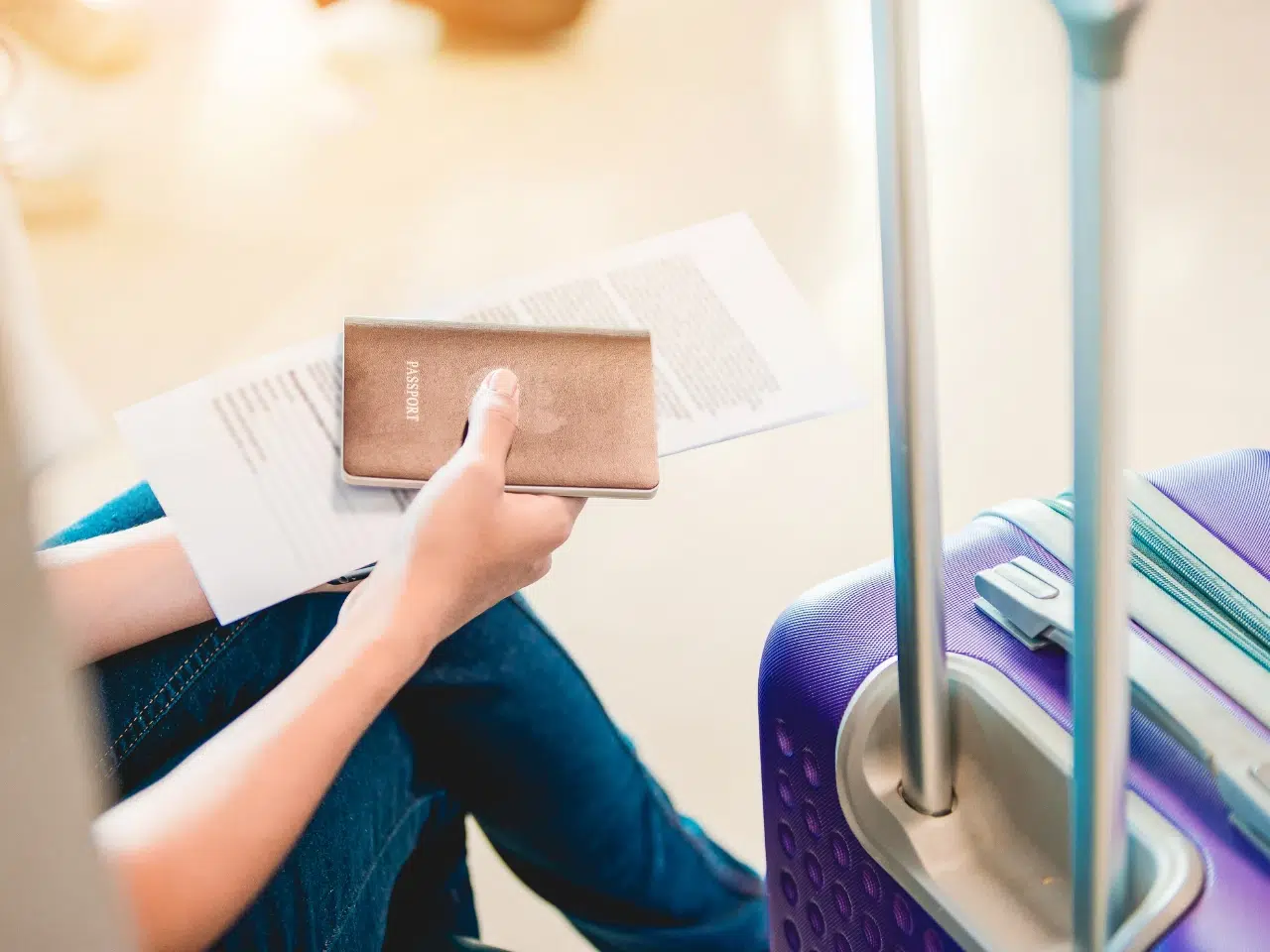 A person sitting with a passport and travel documents in hand next to a purple hard-shell suitcase, preparing for a trip.