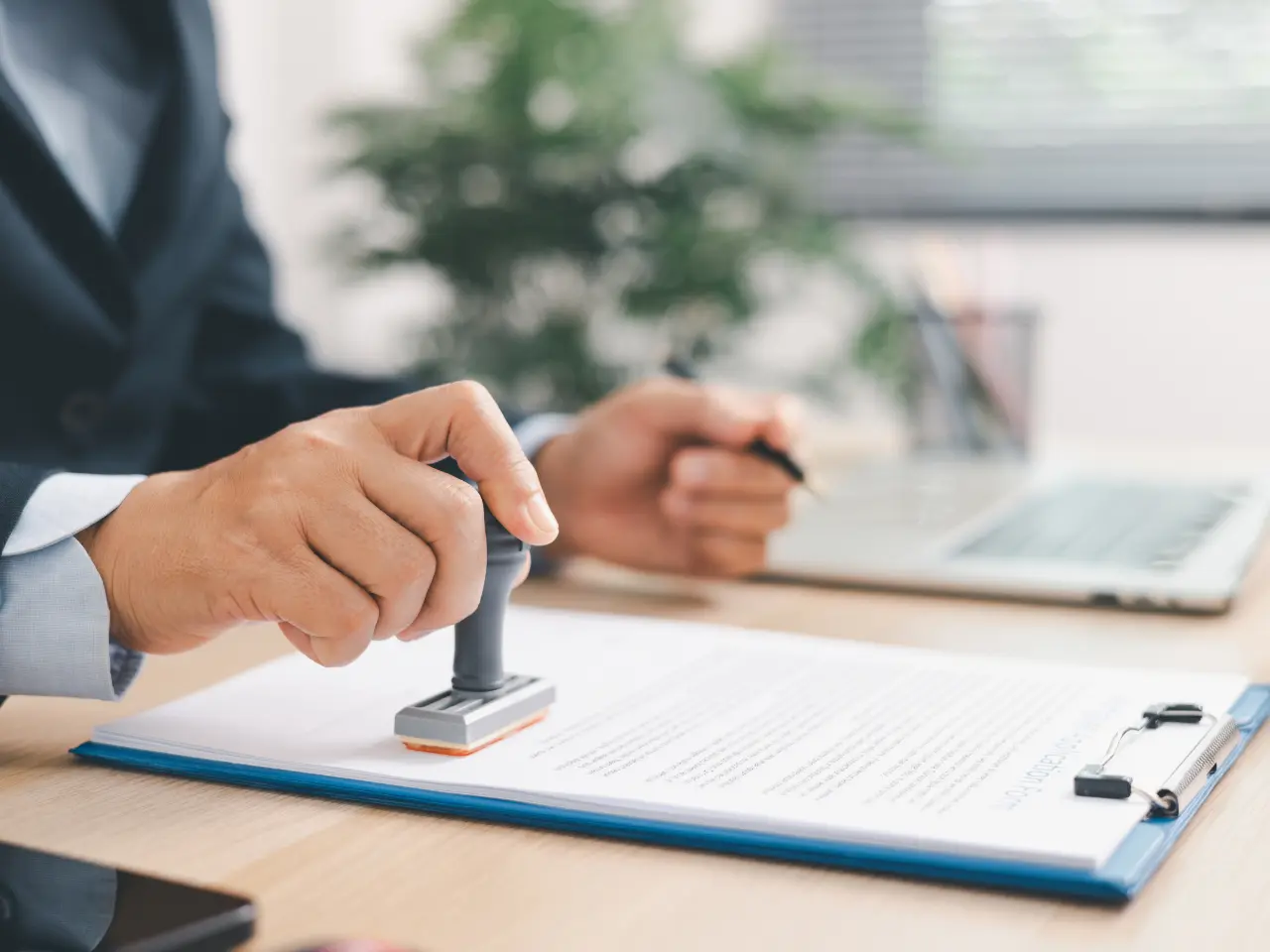 A person in business attire uses a stamp on a document clipped to a blue clipboard, with a pen in the other hand and a laptop and plant visible in the blurred background.