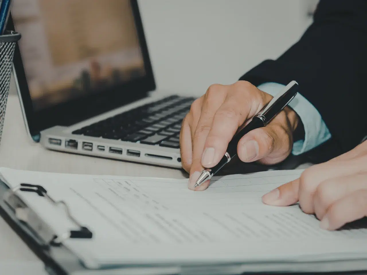 A person in business attire fills out a form on a clipboard with a pen, next to an open laptop on a desk.