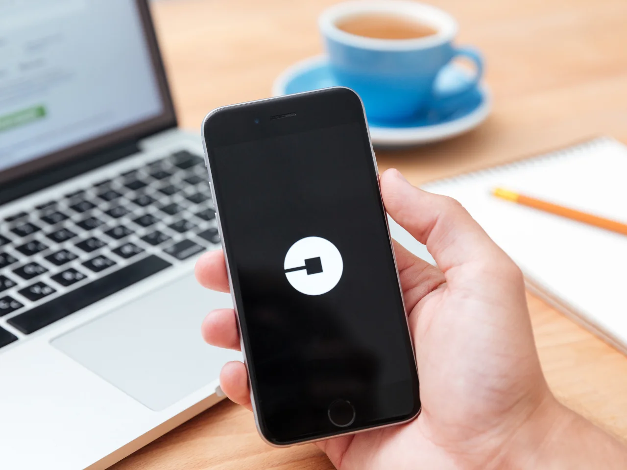 A hand holds a smartphone displaying the Uber app logo. In the background, there is a laptop, a blue cup and saucer, a notepad, and an orange pencil on a wooden table.