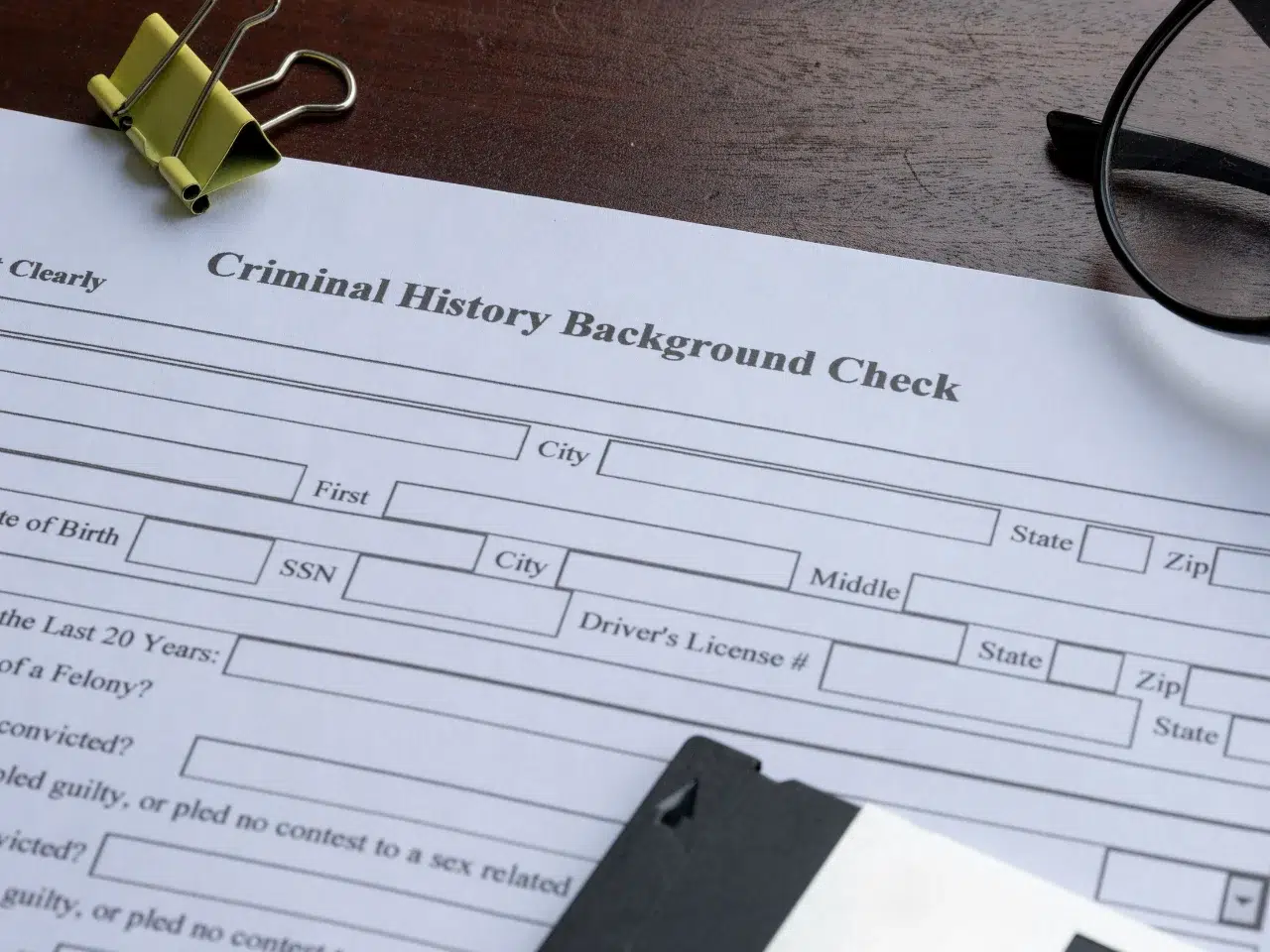 A close-up of a criminal history background check form on a desk with a binder clip, part of a pair of glasses, and a black plastic item partially visible. The form includes spaces for personal information.