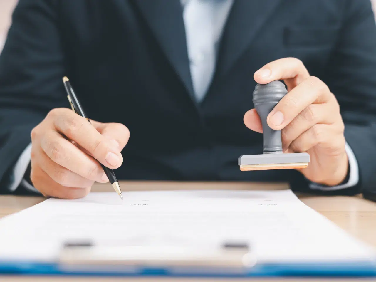 A person in a suit is holding a stamp in one hand and a pen in the other, preparing to stamp and sign a document on a desk.