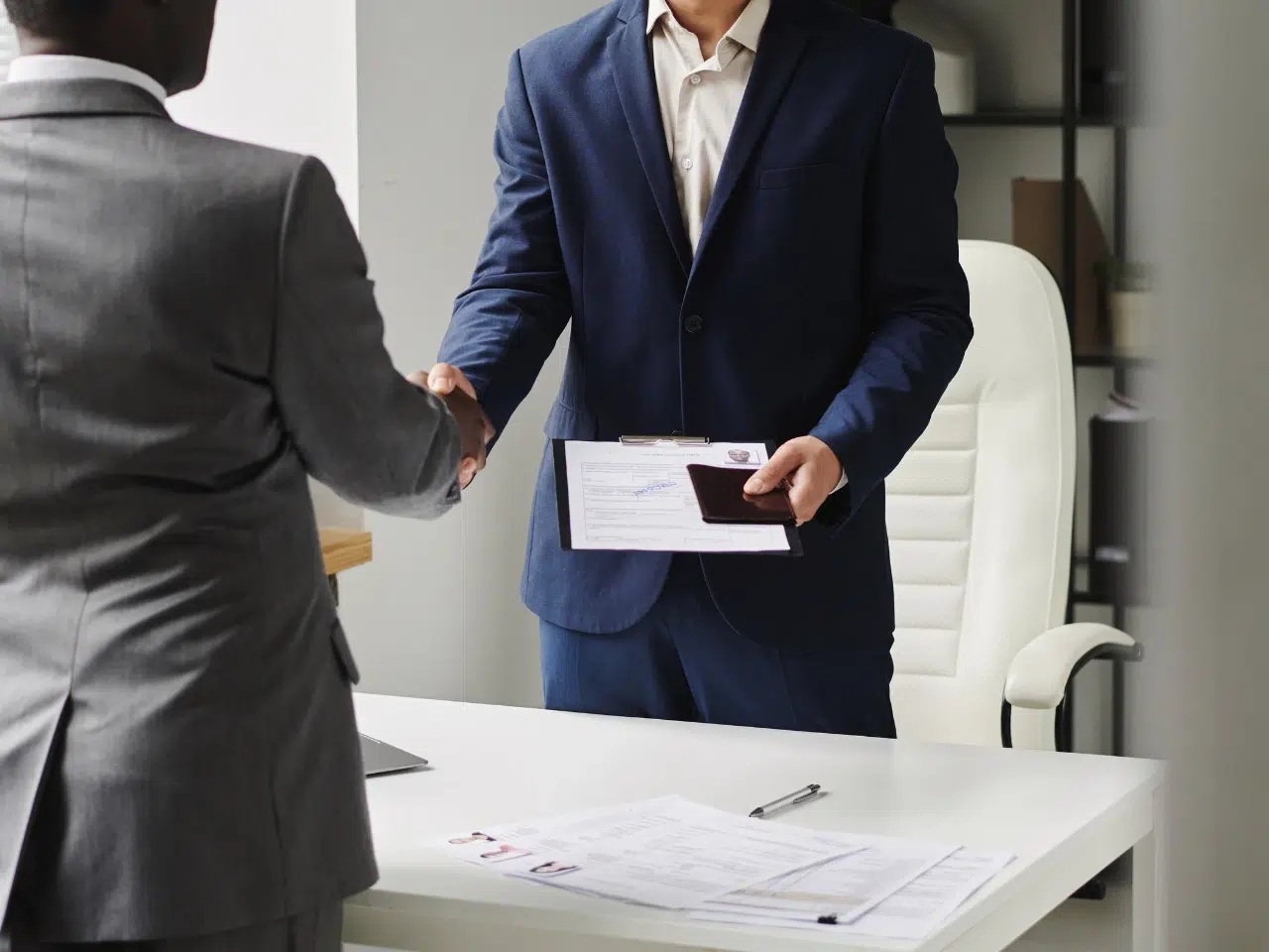 Two men in suits shaking hands in an office. One holds documents and a passport, while the other’s back is to the camera. A desk with papers and a pen is in the foreground.
