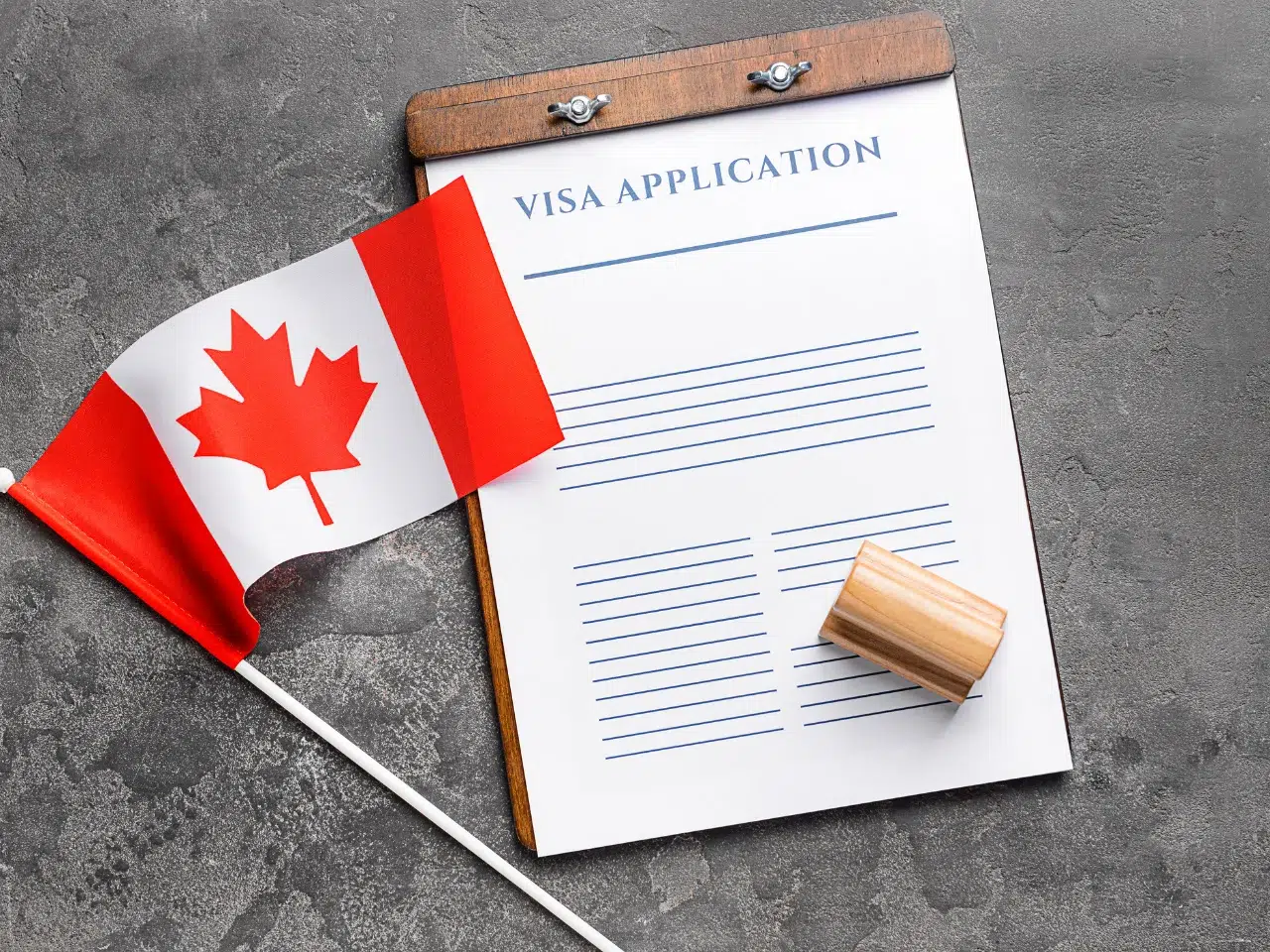 A Canadian flag, a wooden rubber stamp, and a visa application form on a clipboard are arranged on a gray textured surface.
