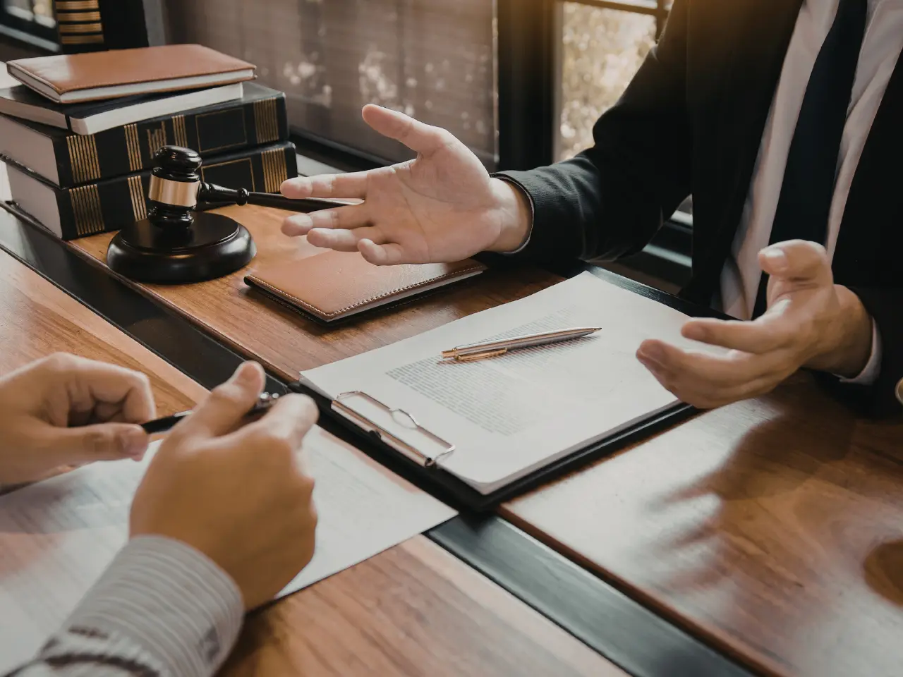 Two people in business attire sit at a desk with documents, a clipboard, a gavel, and books, discussing legal or business matters. One person gestures with their hand while the other holds a pen.